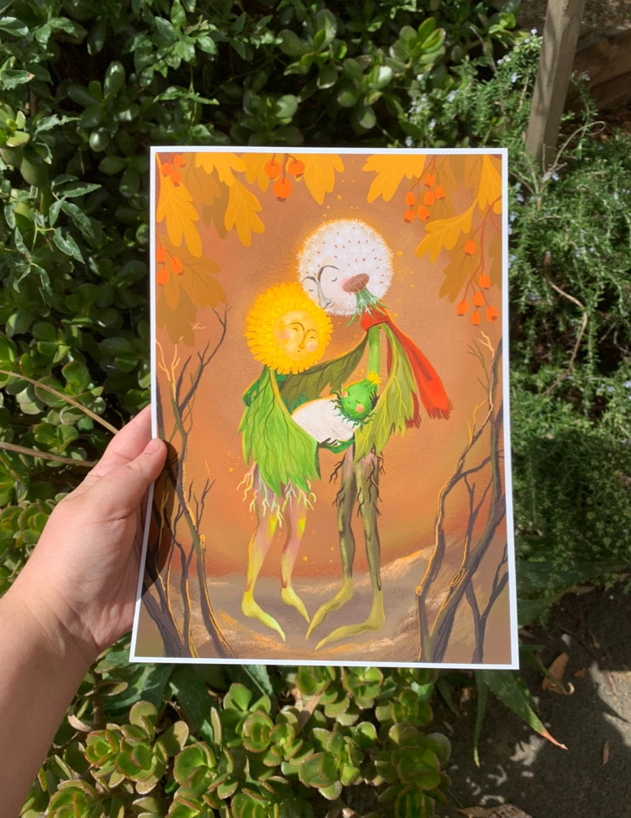 Dandelion Family print held in a hand outdoors against green garden shrubs, showing the dandelion couple and their baby bud