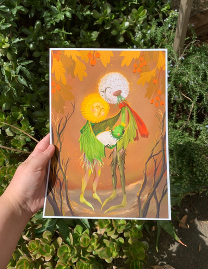 Dandelion Family print held in a hand outdoors against green garden shrubs, showing the dandelion couple and their baby bud