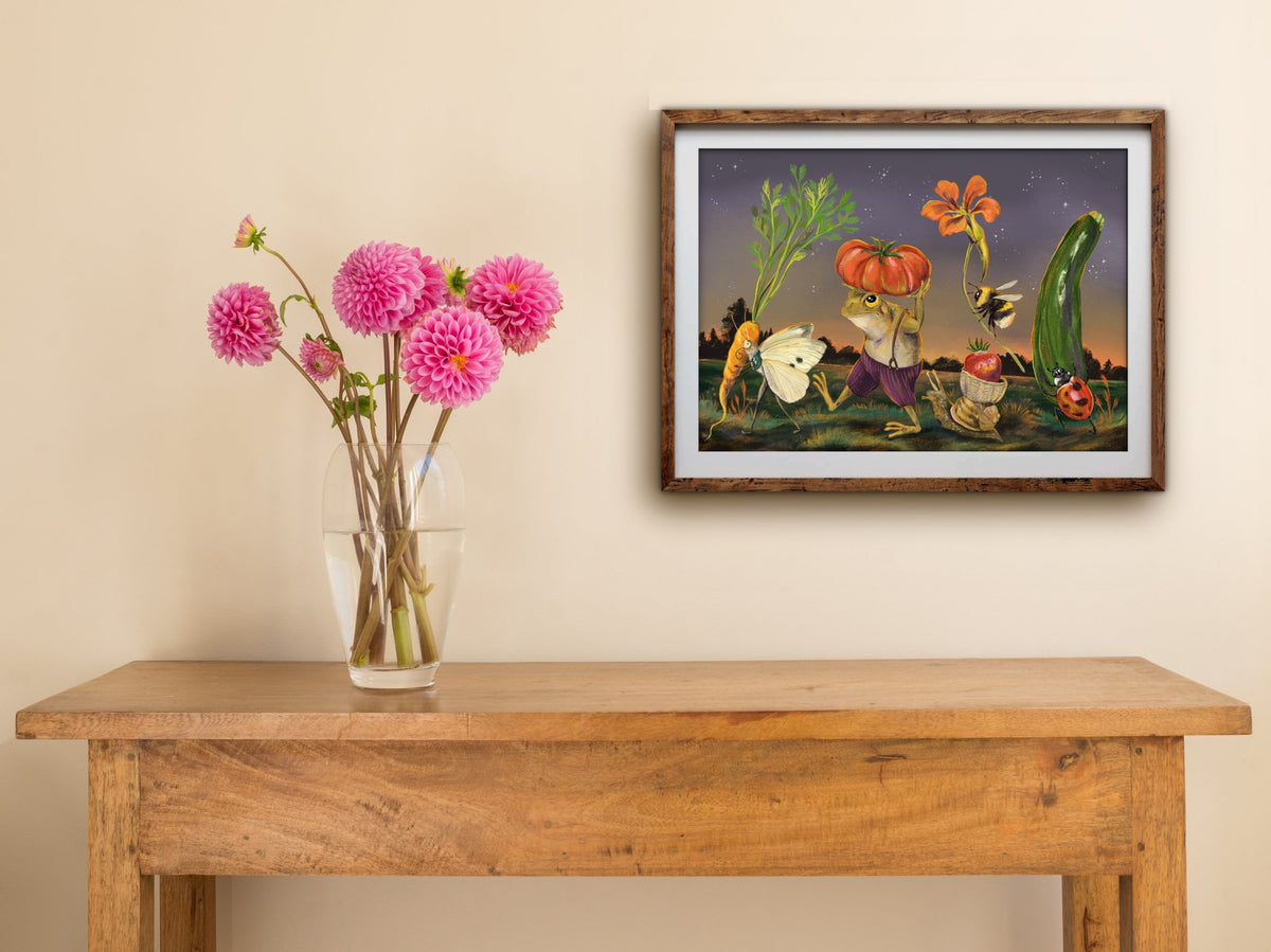 Framed Dawn Harvest print of a frog and garden bugs carrying vegetables at twilight, hung above a wooden table with dahlias