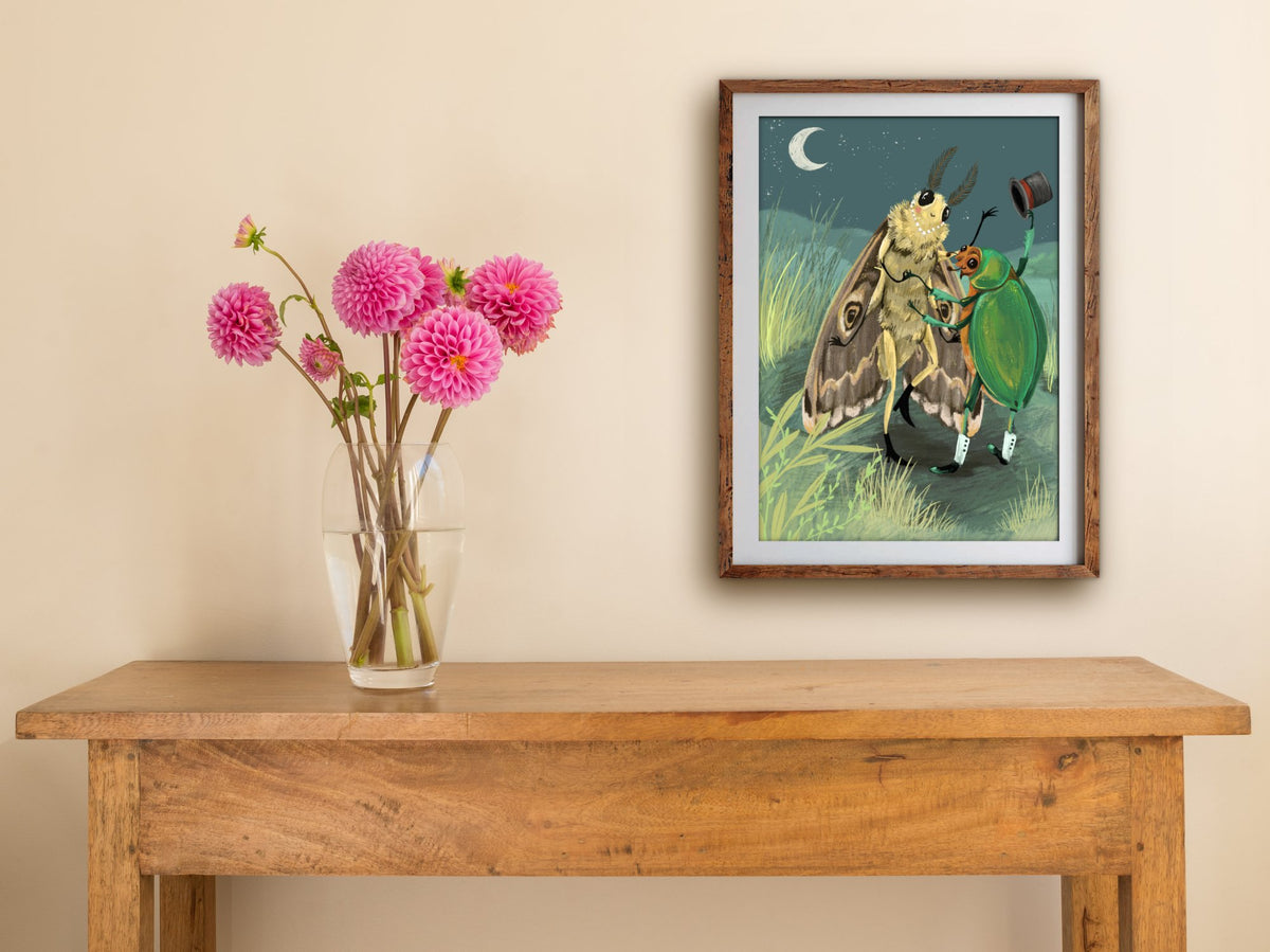 Framed Fancy Dancing print of a moth and beetle dancing under the moon, hung above a wooden side table with pink dahlias