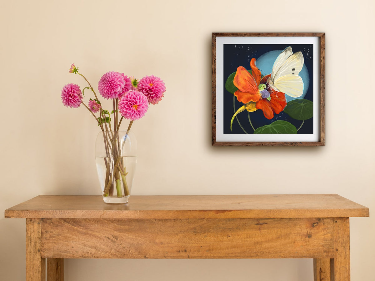Framed Flowerbed print of a butterfly tucking a caterpillar into an orange flower, hung above a wooden table with dahlias