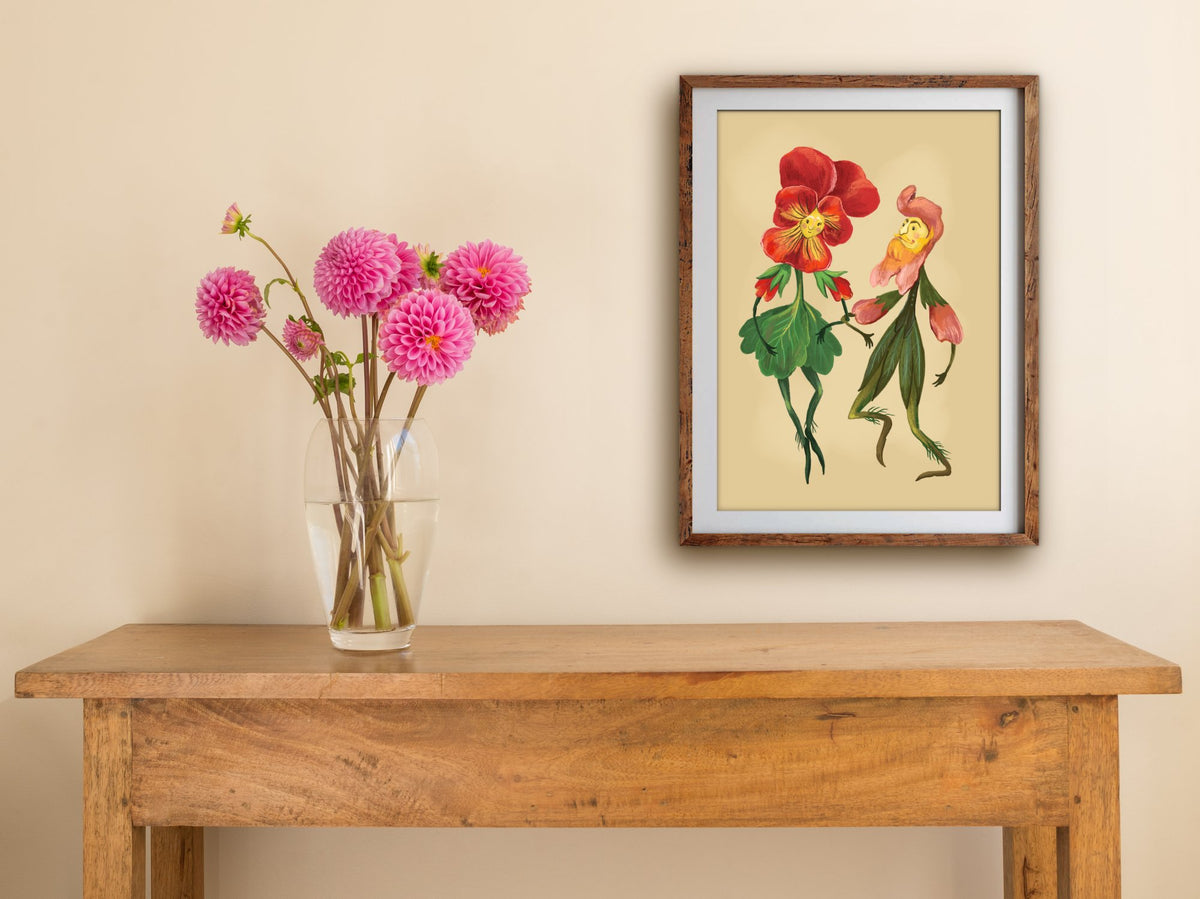 Framed Flower Couple print of a red pansy and pink snapdragon dancing, hung above a wooden side table with pink dahlias