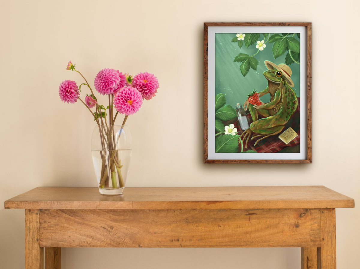 Framed Froggie Picnic print of a frog enjoying a strawberry on a picnic rug, hung above a wooden side table with pink dahlias