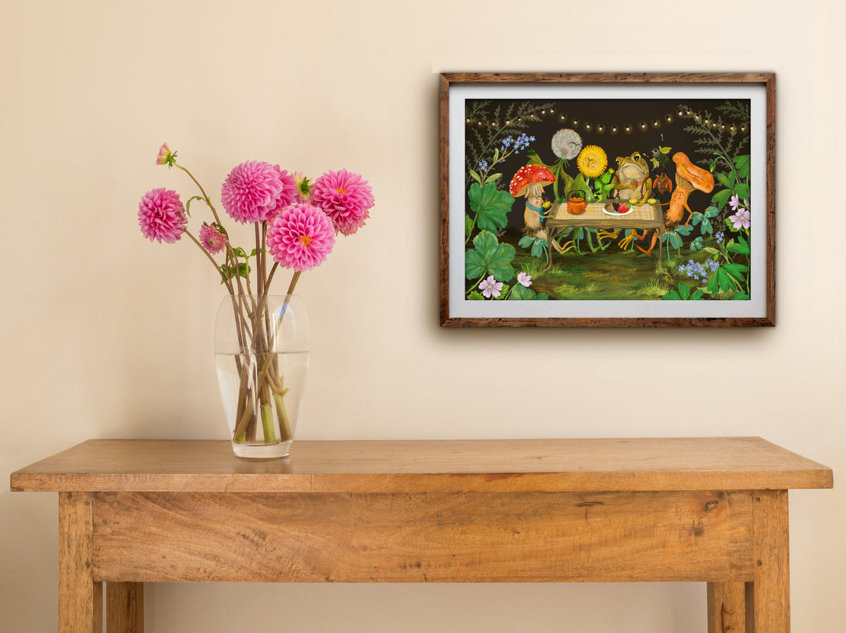 Framed Garden Party print of a toad and mushroom friends sharing tea under string lights, hung above a table with dahlias