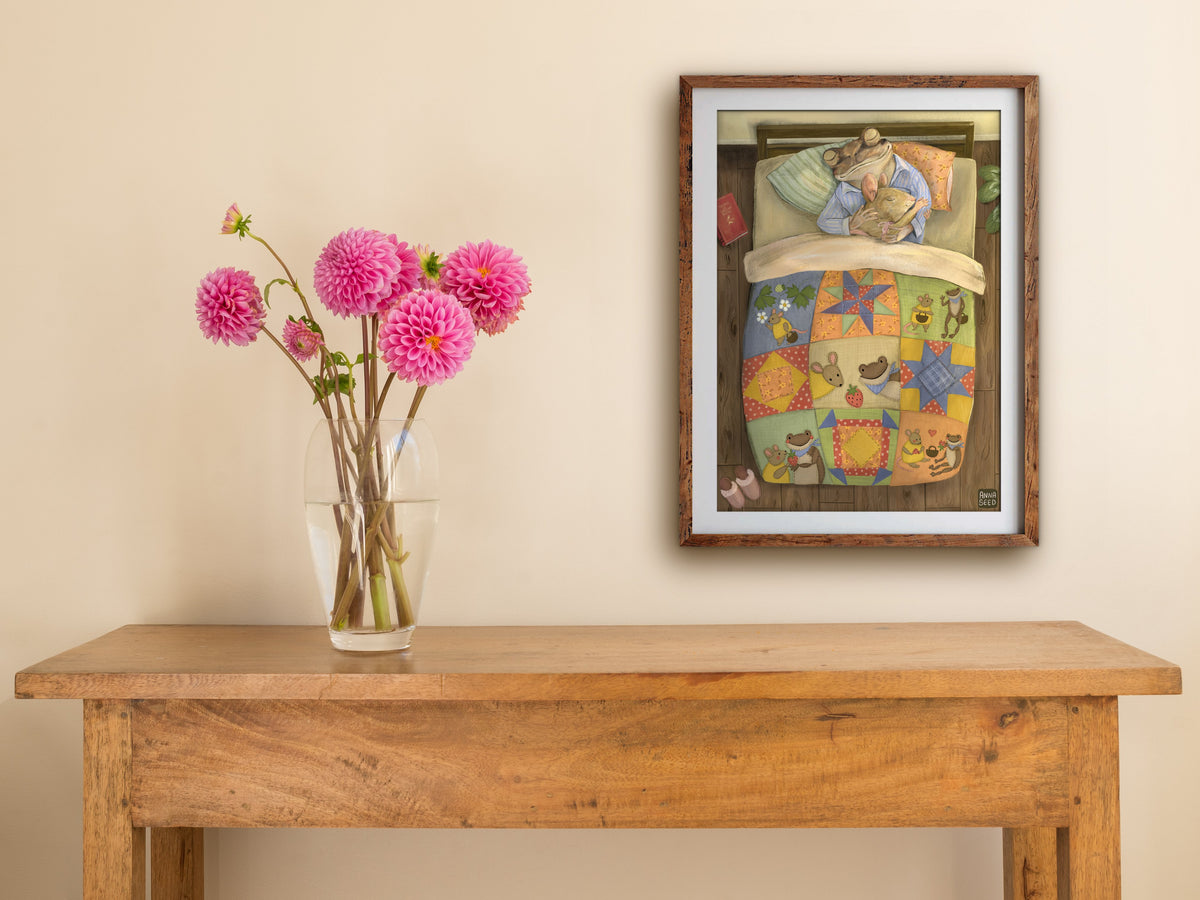Framed Good Morning print of a frog and mouse in bed, hung on a cream wall above a wooden table with pink dahlias