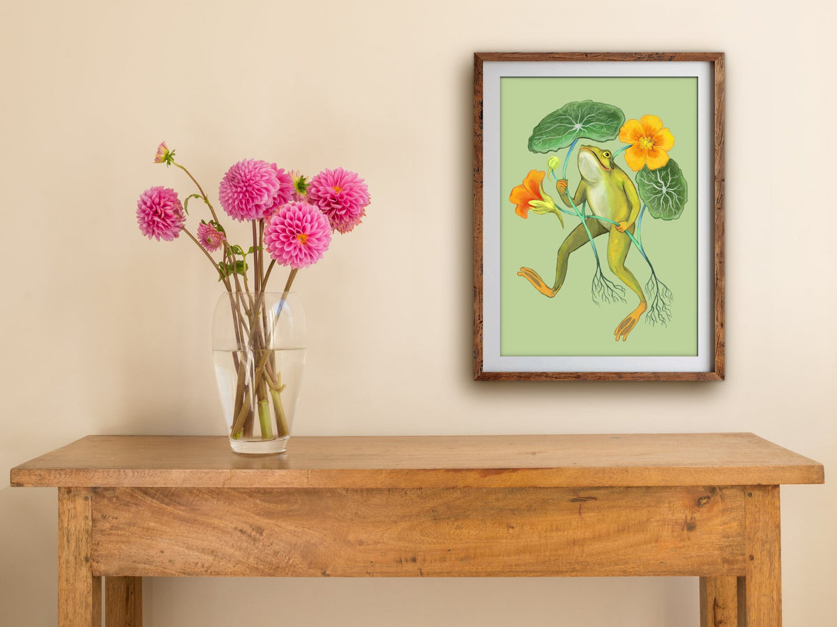 Framed Happy Frog print hanging on a cream wall above a wooden side table with a glass vase of pink dahlias