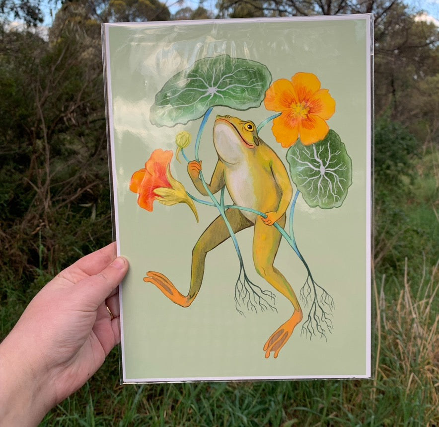 Happy Frog print in a clear sleeve held in a hand outdoors, showing the frog with nasturtiums against green bushland