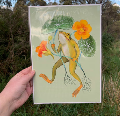 Happy Frog print in a clear sleeve held in a hand outdoors, showing the frog with nasturtiums against green bushland