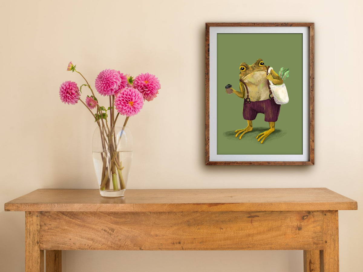Framed Hipster Toad print on a cream wall above a wooden side table with a glass vase of pink dahlias
