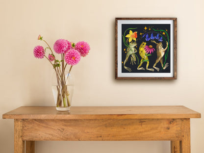 Framed Jolly Frogs print on a cream wall above a wooden side table with a glass vase of pink dahlias