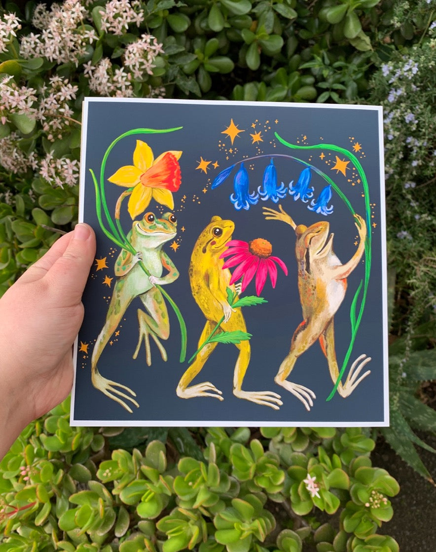 Jolly Frogs print held in a hand over garden succulents, showing three frogs parading with flowers under gold stars