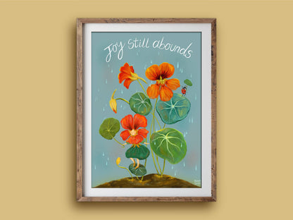 Joy Still Abounds print in a timber frame: orange nasturtiums and round leaves under falling rain, with a tiny ladybird