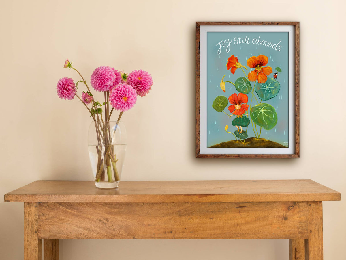 Framed Joy Still Abounds print on a cream wall above a wooden side table with a glass vase of pink dahlias