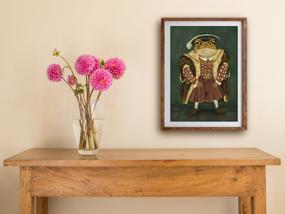 Framed King Toad portrait print on a cream wall above a wooden side table with a glass vase of pink dahlias