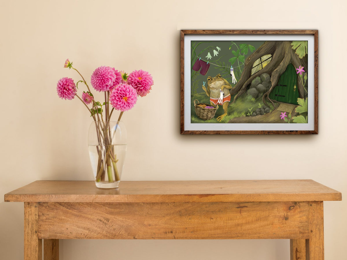 Framed landscape Laundry Toad print on a cream wall above a wooden side table with a glass vase of pink dahlias