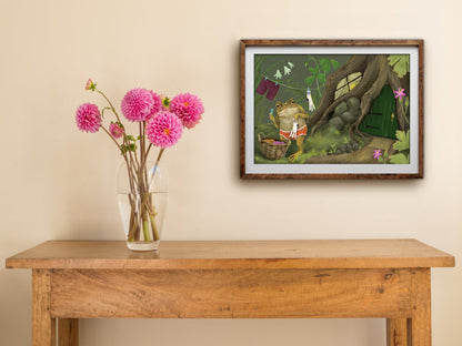 Framed landscape Laundry Toad print on a cream wall above a wooden side table with a glass vase of pink dahlias