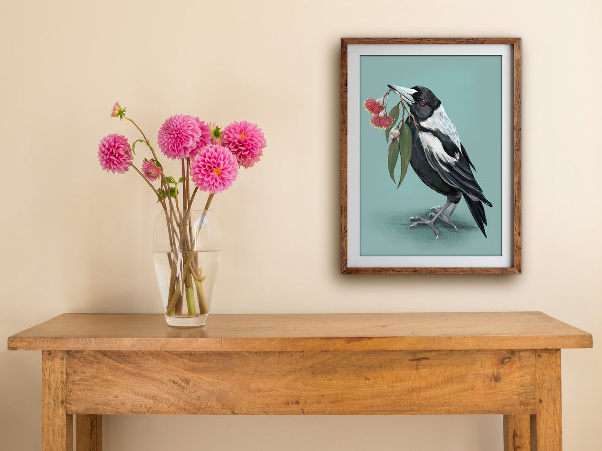 Framed Magpie print on a cream wall above a wooden side table with a glass vase of pink dahlias