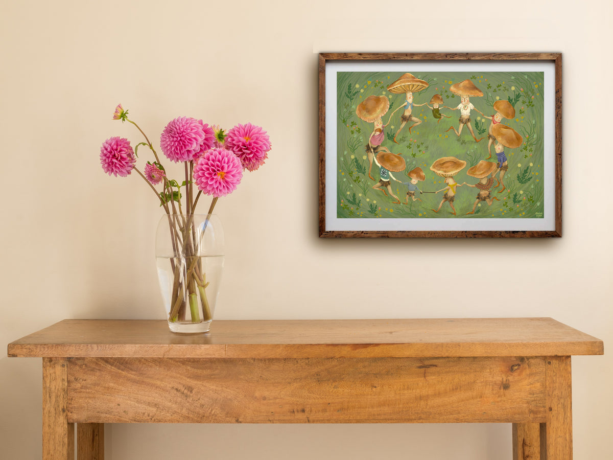 Framed landscape Merry Dance print on a cream wall above a wooden side table with a glass vase of pink dahlias