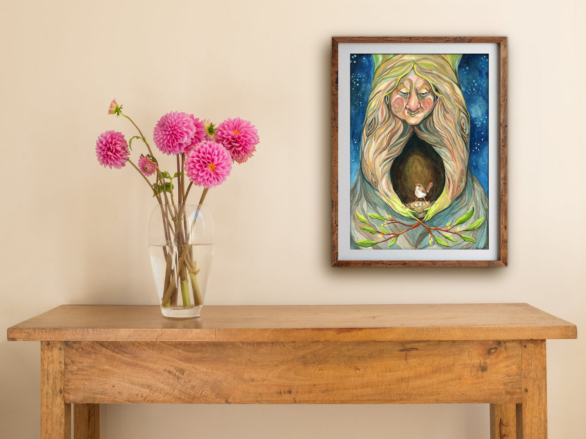 Framed Mother Nature print hung above a wooden table with pink dahlias, showing a tree spirit and wren on starry blue