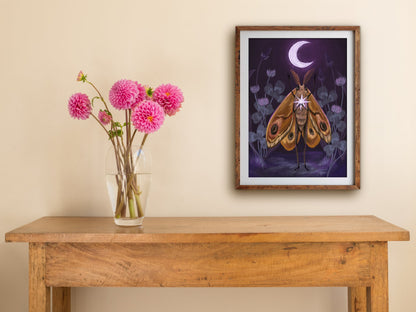 Framed Moth's Dream print hung above a wooden table with pink dahlias, its moth and crescent moon on violet