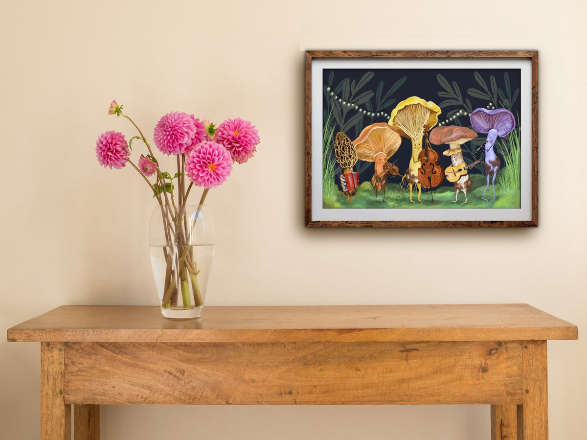 Framed Mushroom Band print hung above a wooden table with pink dahlias, mushroom musicians playing on a mossy stage