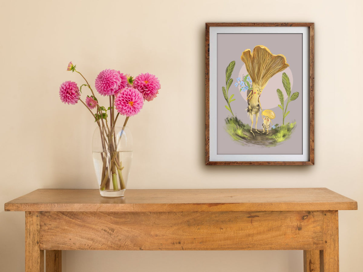 Framed Mushroom Mama print hung above a wooden table with pink dahlias, a mushroom mother and child among leaves