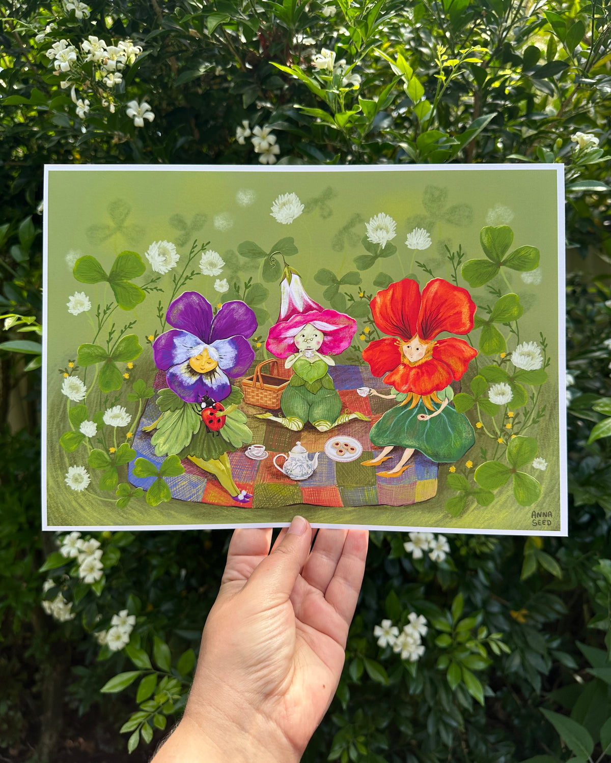 Petal Picnic print held up in a hand outdoors before a hedge of white flowers, three flower friends picnicking in clover