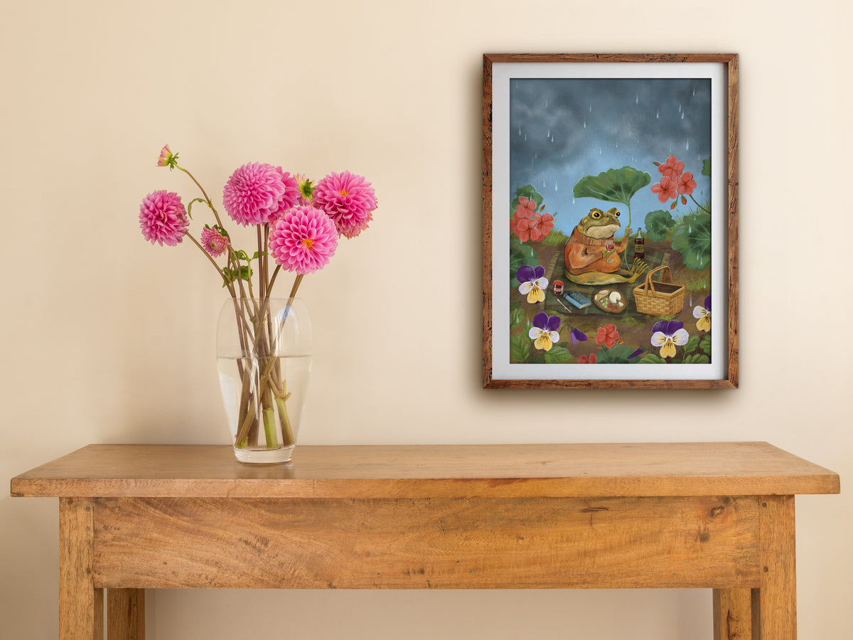 Framed Picnic Toad print hung above a wooden table with pink dahlias, a rained-on toad picnicking among geraniums