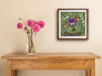 Framed Pretty Miss Pansy print hung above a wooden table with pink dahlias, a dancing pansy among orange butterflies