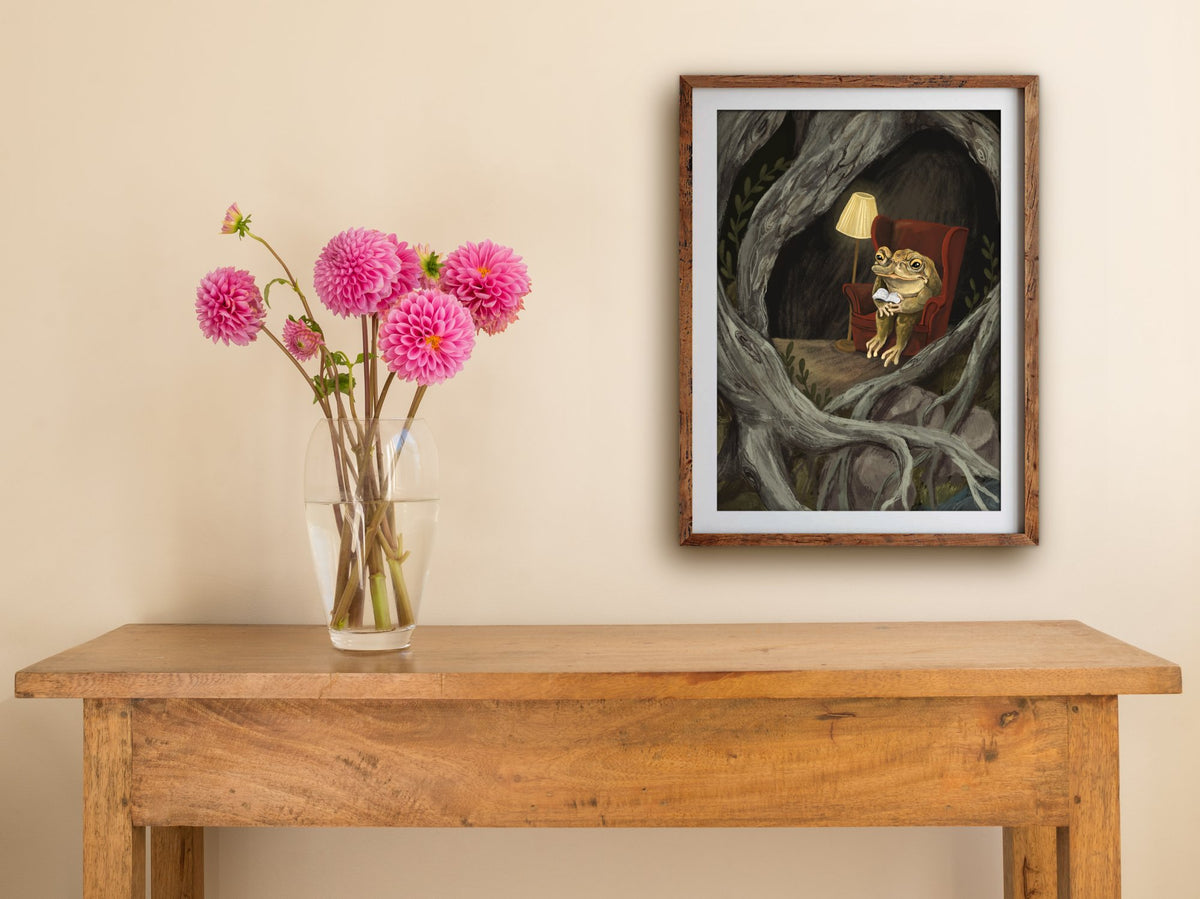 Framed Reading Toad print hung above a wooden table with pink dahlias, a toad reading in a red wingback chair