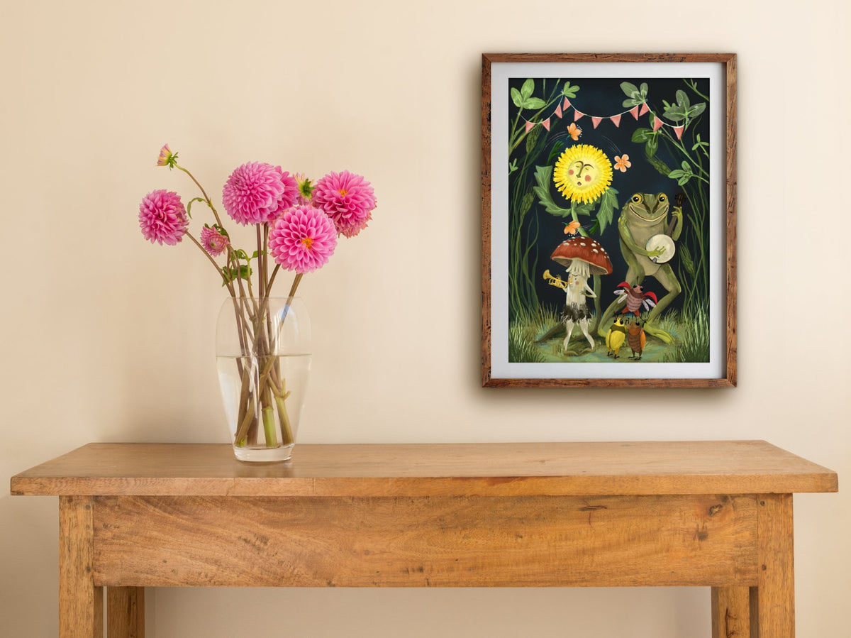 Framed Showtime print hung above a wooden table with pink dahlias, a banjo frog and mushroom trumpeter celebrating spring