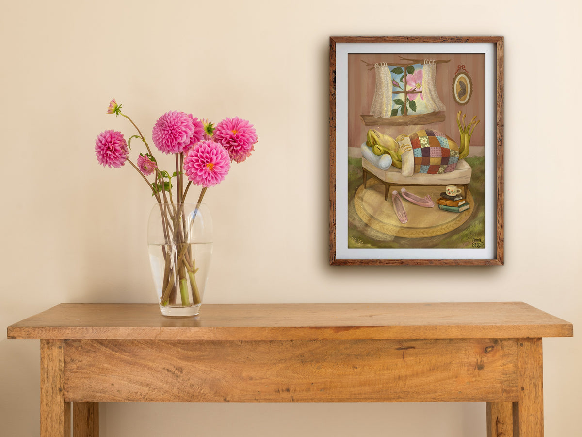 Framed Sleeping In print hung above a wooden table with pink dahlias, showing a frog snoozing under a patchwork quilt