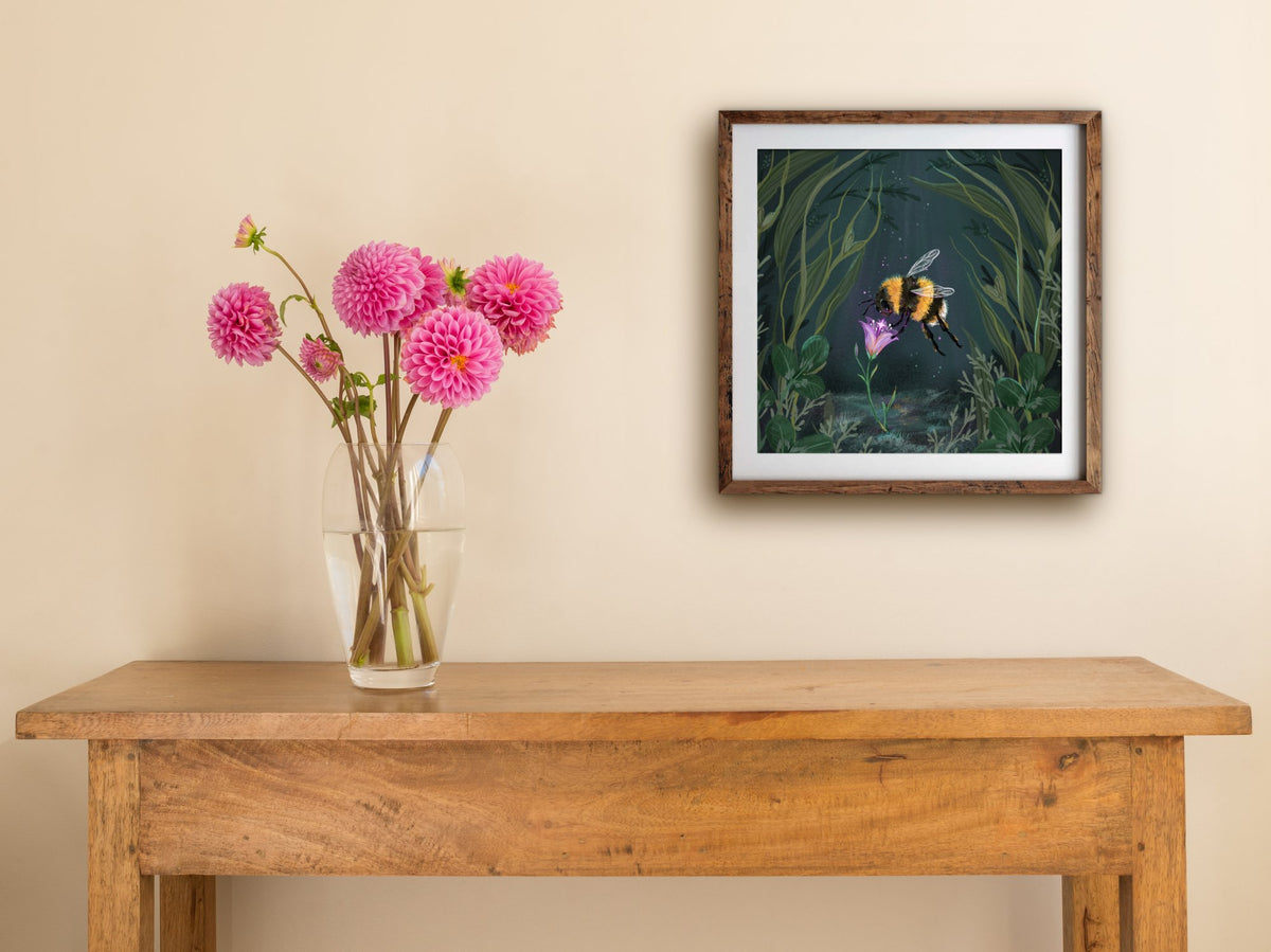Framed Starflower square print hung above a wooden table with pink dahlias, showing the bee and glowing flower
