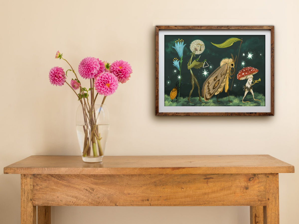 Framed Starlight Parade landscape print hung above a wooden table with pink dahlias, showing the night-time parade