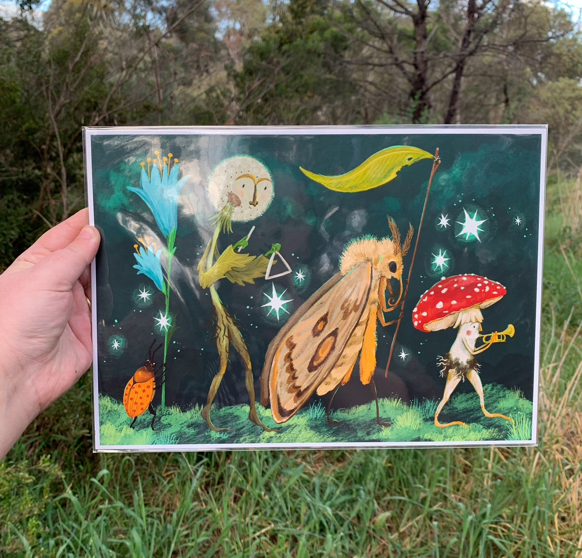 Hand holding the Starlight Parade print outdoors against bushland, its moth, dandelion and toadstool band glowing