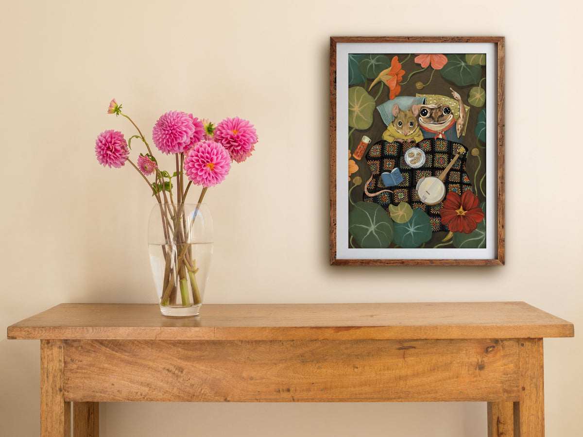 Framed Starry Eyes print hung above a wooden table with pink dahlias, showing the cosy mouse and frog under a blanket