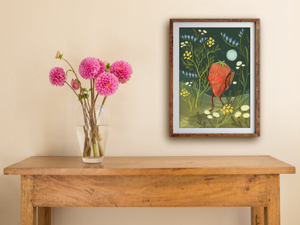 Framed Strawberry Adventurer print hung above a wooden table with pink dahlias, the strawberry among blue and yellow blooms