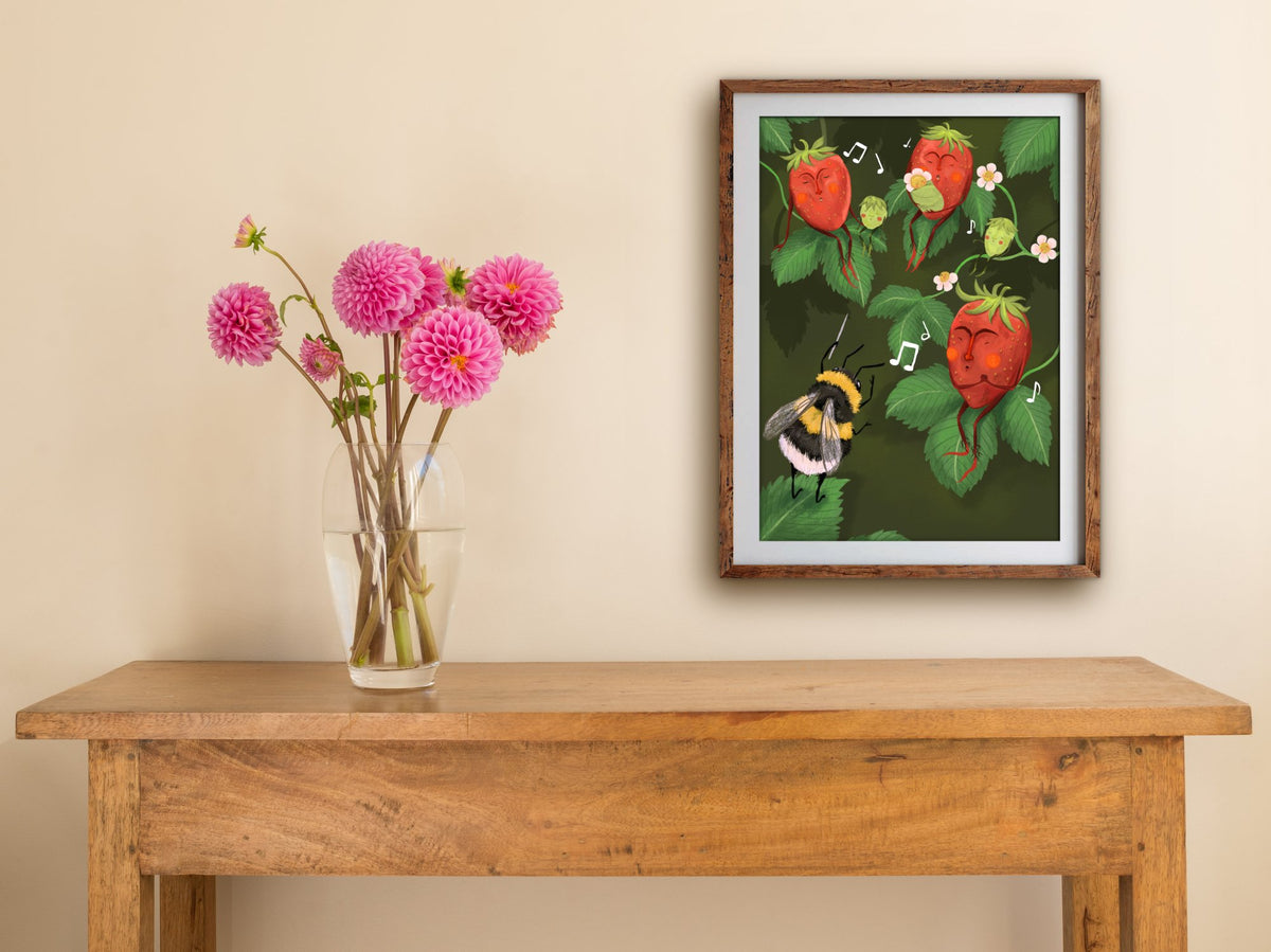 Framed Strawberry Choir print hung above a wooden table with pink dahlias, showing the bee conducting a strawberry choir