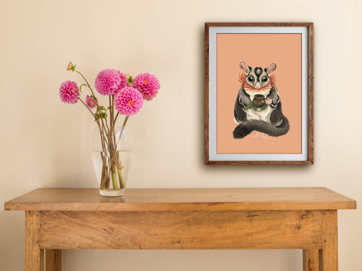 Framed Sugar Glider Teatime print hung above a wooden table with pink dahlias, the possum's tea time on a peach background
