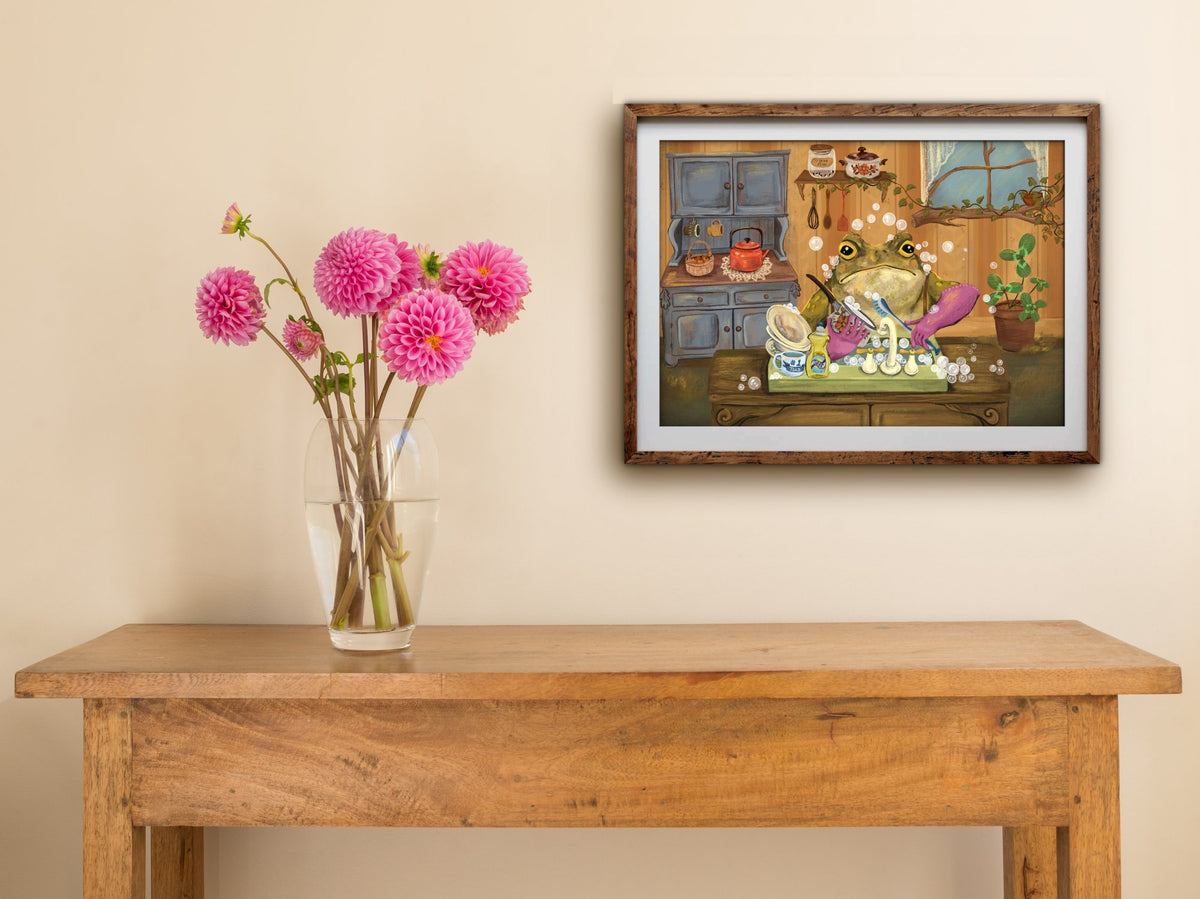 Framed Toady Dishes landscape print hung above a wooden table with pink dahlias, the toad washing up among soap bubbles