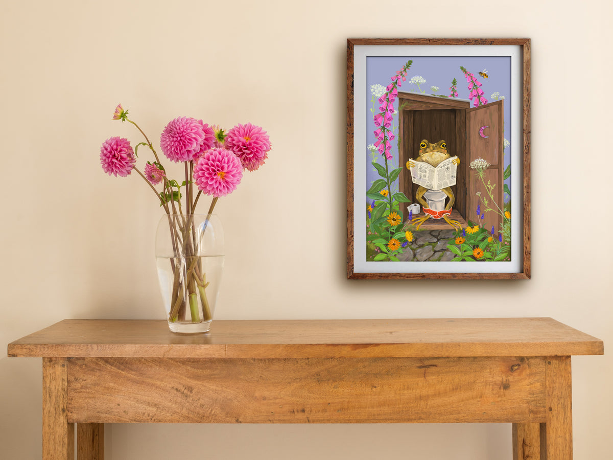 Framed Toilet Toad print hung above a wooden table with pink dahlias, showing the toad in his flower-framed outhouse