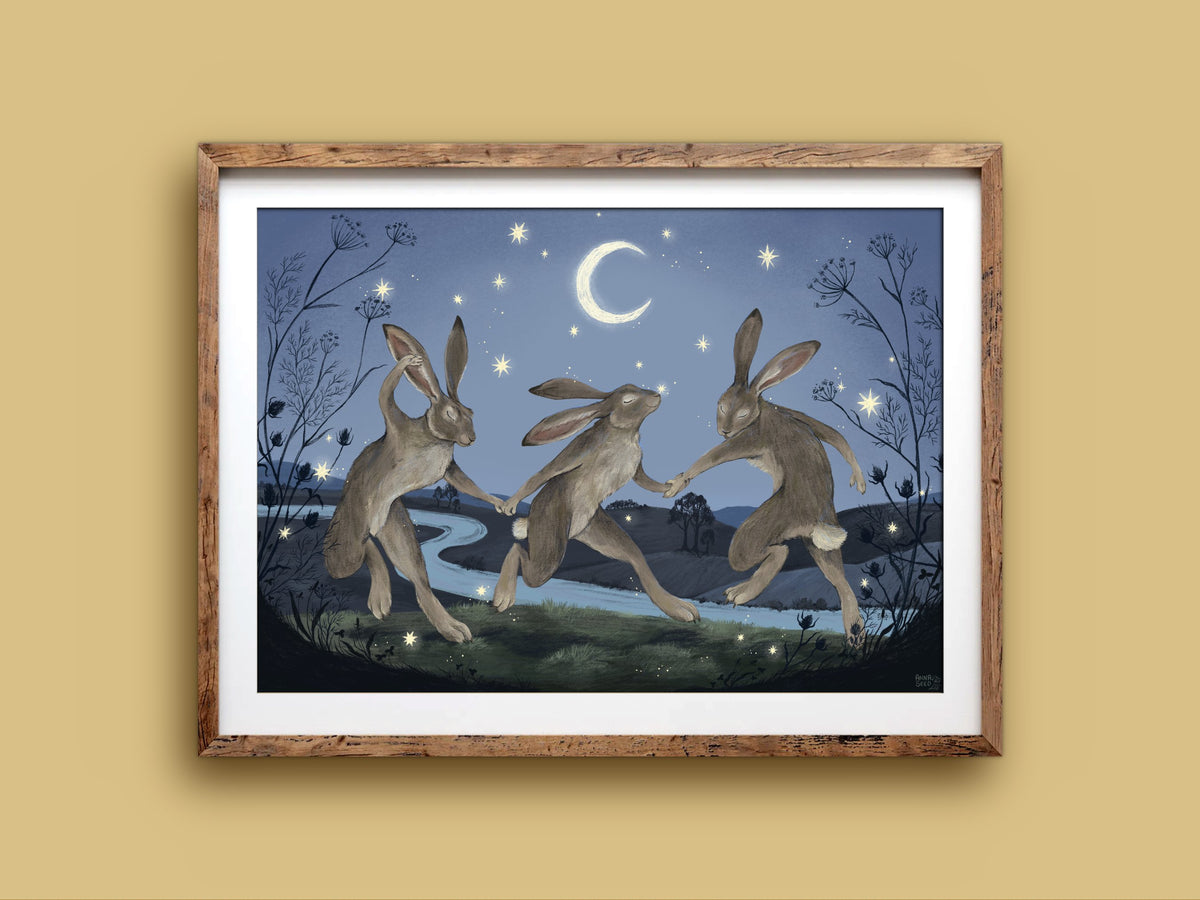 Velveteen Dancers landscape print in a rustic timber frame on a mustard wall, three hares dancing in blue moonlight