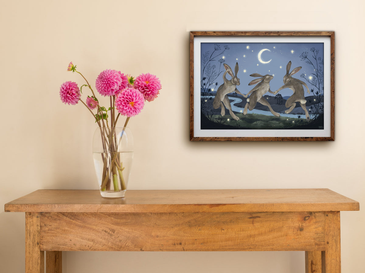 Framed Velveteen Dancers print hanging above a wooden side table with a vase of pink dahlias against a cream wall