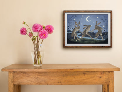 Framed Velveteen Dancers print hanging above a wooden side table with a vase of pink dahlias against a cream wall