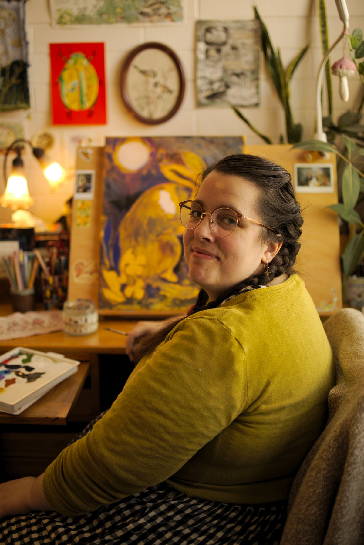 Anna Seed smiling at her desk in a mustard cardigan, in front of a bright yellow painting and her art-covered wall