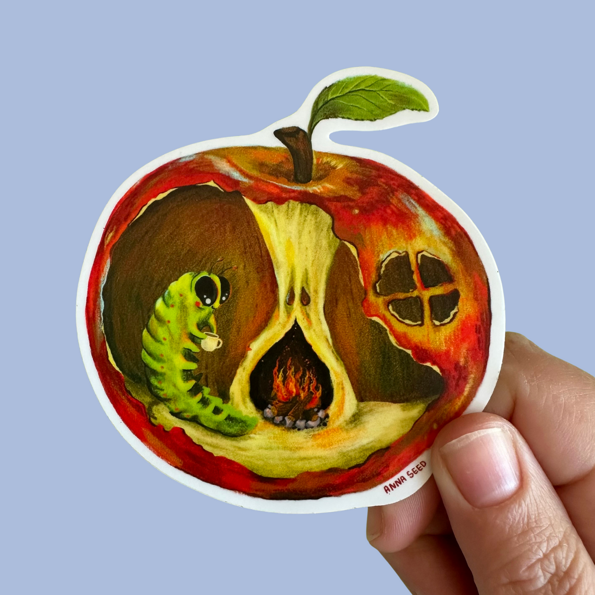 Apple Cottage sticker by Anna Seed: a green grub sipping tea by a fireplace inside a red apple house, held on blue