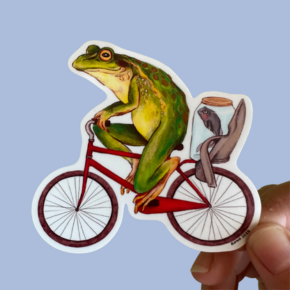 Bicycle Frog sticker by Anna Seed: a green frog riding a red bike with a tadpole in a jar in the child seat, on blue