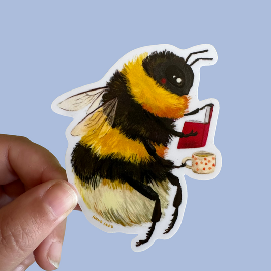 Bumblebee sticker by Anna Seed: a fluffy bumblebee reading a red book and holding a spotty teacup, held on blue