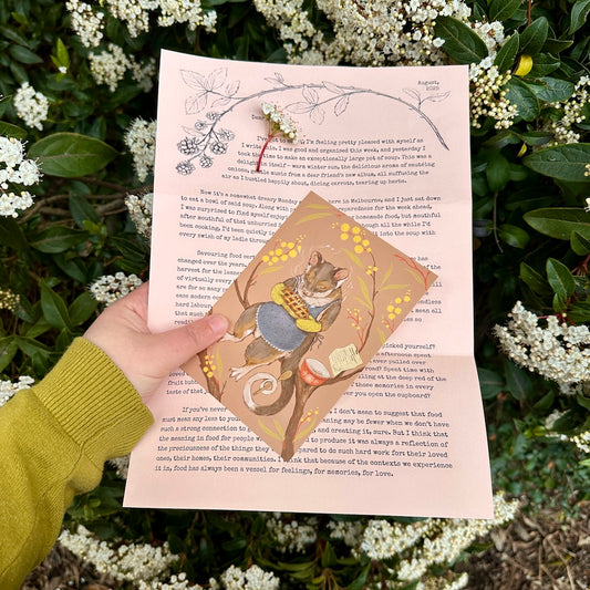 Whimsy Way letter #2 by Anna Seed on pink paper, held with a possum art card in front of white-flowering shrubs