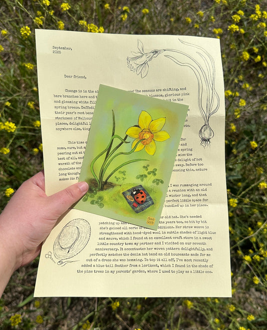 Whimsy Way letter #3 by Anna Seed, held with a daffodil and ladybird art card in front of yellow wildflowers